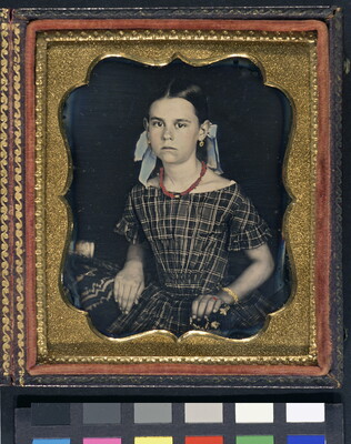 A hand-colored photograph in a decorative gold frame of a young White girl seated, wearing a plaid dress and bows in her hair.