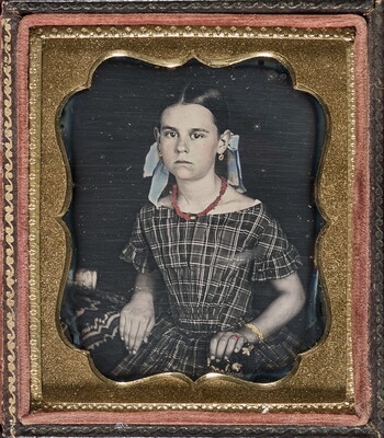 A hand-colored photograph in a decorative gold frame of a young White girl seated, wearing a plaid dress and bows in her hair.