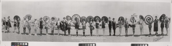 A black-and-white panoramic photograph of a group of women, lined side-by-side on a beach, wearing elaborate bathing costumes, hats, and holding parasols.