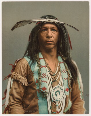 A colored photograph portrait of an Indigenous man in traditional clothing, including beaded necklaces and large feathers on his head.