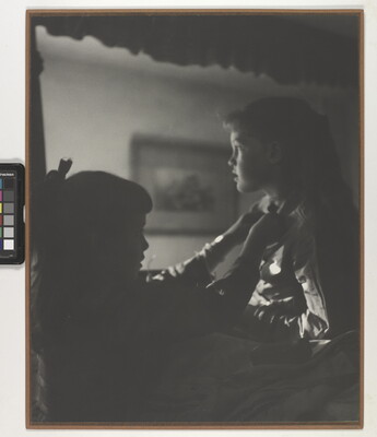 A black-and-white photograph of two young White girls, one fastening a ribbon around the neck of the other, both in shadow.