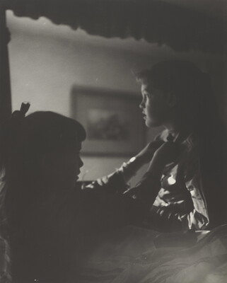 A black-and-white photograph of two young White girls, one fastening a ribbon around the neck of the other, both in shadow.