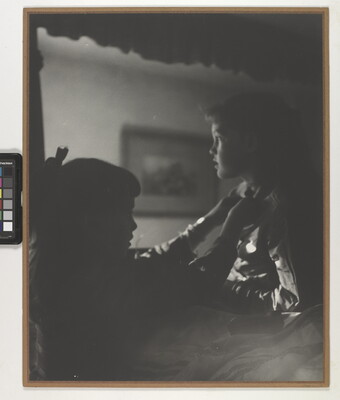 A black-and-white photograph of two young White girls, one fastening a ribbon around the neck of the other, both in shadow.
