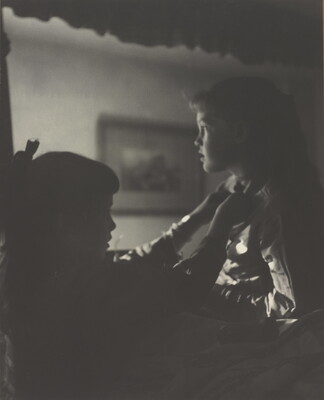 A black-and-white photograph of two young White girls, one fastening a ribbon around the neck of the other, both in shadow.
