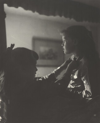 A black-and-white photograph of two young White girls, one fastening a ribbon around the neck of the other, both in shadow.