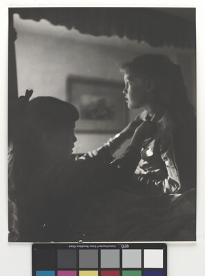A black-and-white photograph of two young White girls, one fastening a ribbon around the neck of the other, both in shadow.