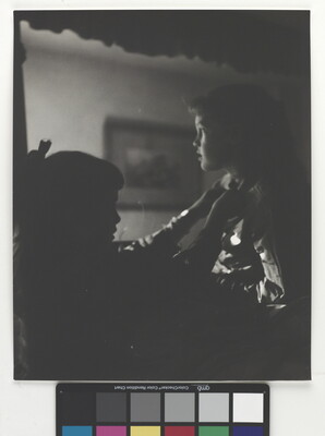 A black-and-white photograph of two young White girls, one fastening a ribbon around the neck of the other, both in shadow.