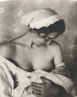 A black-and-white photograph of a White woman in a nightcap and white flowing blouse nursing an infant.