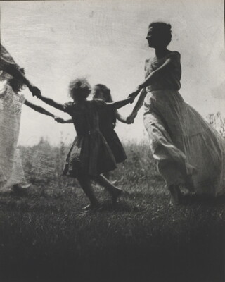 A black-and-white photograph of a White woman and children wearing long dresses holding hands to form a circle in a grassy field.