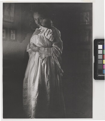A black-and-white photograph of a standing White woman holding a baby wearing a long white nightgown.