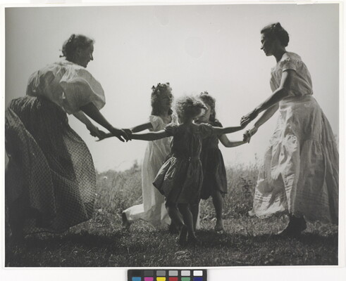 A black-and-white photograph of two White women and three White children outside, holding hands and dancing in a circle.