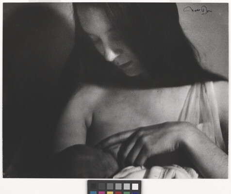 A black-and-white photograph of a White woman with long, dark hair breastfeeding a baby.