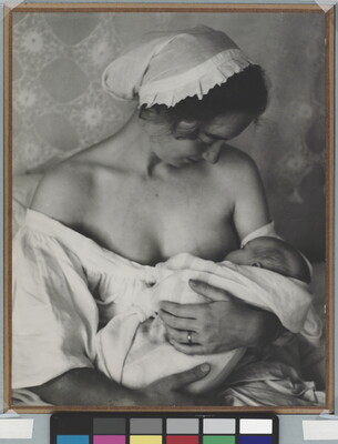 A black-and-white photograph of a White woman in a nightcap and white flowing blouse nursing an infant.