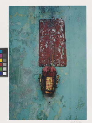A color photograph of a textured blue wall with a dark red rectangle on which hangs a metallic bucket of incense.