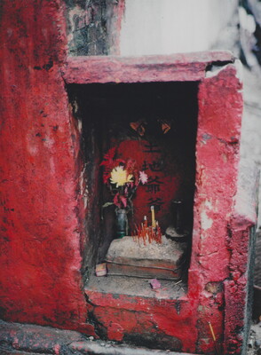 A color photograph of a rectangular recess in a textured red wall housing flowers and incense.