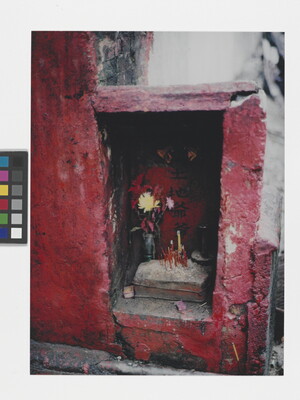 A color photograph of a rectangular recess in a textured red wall housing flowers and incense.