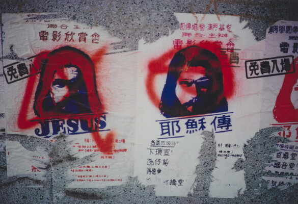 A color photograph of torn flyers printed with images of Jesus on what looks like a concrete wall; red markings are spray painted over the flyers.