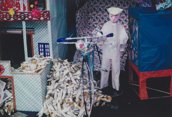 A color photograph of a doll or puppet in a white outfit standing next to a bicycle and a pile of rolled paper packets.