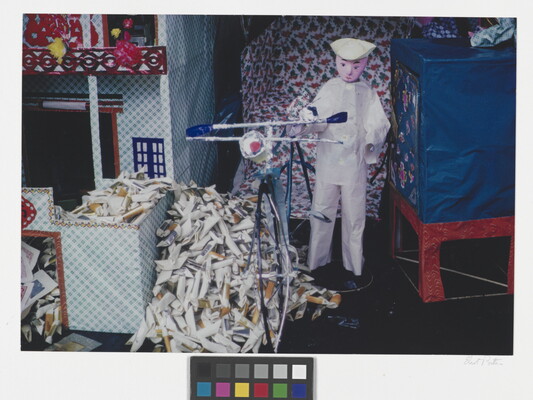 A color photograph of a doll or puppet in a white outfit standing next to a bicycle and a pile of rolled paper packets.