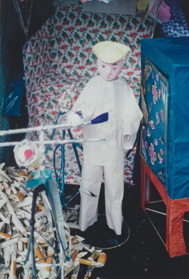 A color photograph of a doll or puppet in a white outfit standing next to a bicycle and a pile of rolled paper packets.