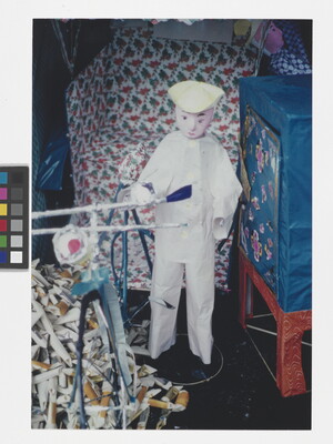 A color photograph of a doll or puppet in a white outfit standing next to a bicycle and a pile of rolled paper packets.