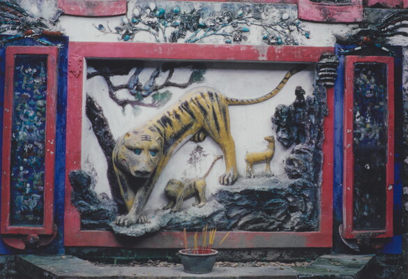 A color photograph of a wall with a relief carving and colorful mural of a large tiger and several smaller animals climbing through trees toward a pot of incense placed in front of the mural.