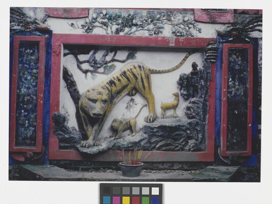 A color photograph of a wall with a relief carving and colorful mural of a large tiger and several smaller animals climbing through trees toward a pot of incense placed in front of the mural.