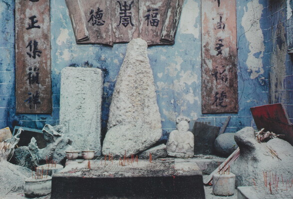 A color photograph of a shrine with blue walls filled with abstract and figurative stone sculptures and incense sticks in pots scattered in front of them.