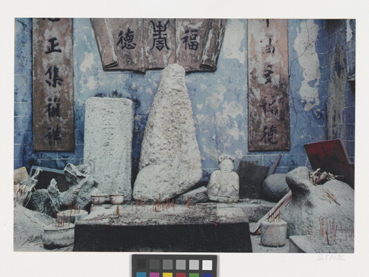 A color photograph of a shrine with blue walls filled with abstract and figurative stone sculptures and incense sticks in pots scattered in front of them.