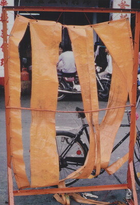 A color photograph of a street with bicycles and motorcycles seen through strips of paper or fabric suspended on a red frame.