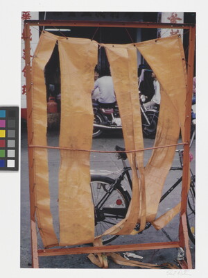 A color photograph of a street with bicycles and motorcycles seen through strips of paper or fabric suspended on a red frame.