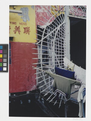A color photograph of a wall painted red and yellow next to a white metal gate on which hangs some laundry.
