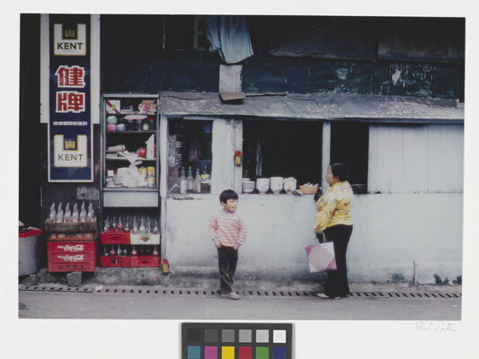 A color photograph of an Asian woman and young child standing at the window of a small white building that looks like it serves food.