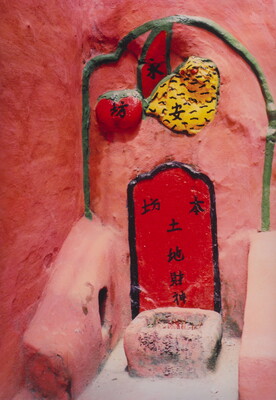 A color photograph of a shrine inset in a red stone wall, with a container for incense and relief sculptures of fruit.
