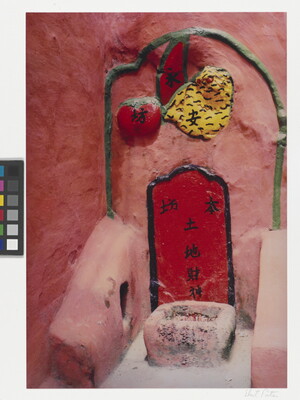 A color photograph of a shrine inset in a red stone wall, with a container for incense and relief sculptures of fruit.