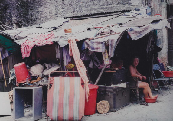 A color photograph of an Asian person sitting beneath a crowded shack with their bare feet in a red bucket.