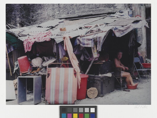 A color photograph of an Asian person sitting beneath a crowded shack with their bare feet in a red bucket.