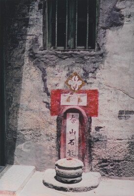 A color photograph of a shrine inset in a plaster wall, red and white geometric details are painted on the wall,  a cylindrical incense burner sits in front of it.