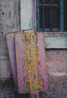 A color photograph of weathered pink and yellow wood signs with Asian characters leaning against a stone wall next to a window.