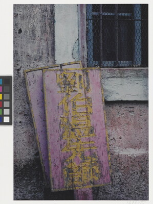 A color photograph of weathered pink and yellow wood signs with Asian characters leaning against a stone wall next to a window.