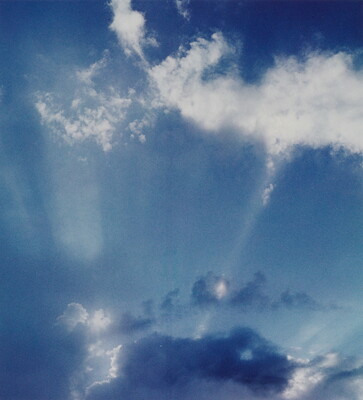 A color photograph of white clouds in a blue sky and sun rays that create white streaks.