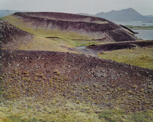 A color photograph of a crater on a rocky and green landscape with water and mountains in the distance.