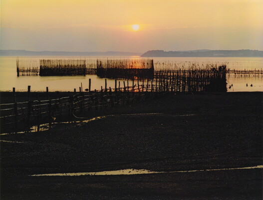A color photograph of a sunset over a bay with a rocky beach and a wooden dock in the foreground.
