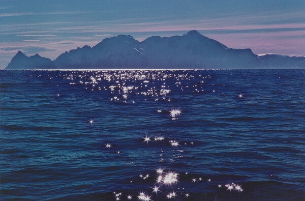 A color photograph of a dark mountain range under a pink sky beyond gentle bright blue waves with light reflecting off of the ripples.