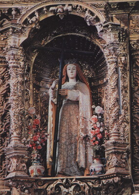 A color photograph of an ornately decorated niche featuring a statue of a woman holding a cross flanked by flower vases.