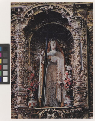 A color photograph of an ornately decorated niche featuring a statue of a woman holding a cross flanked by flower vases.