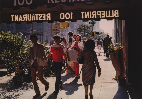A color photograph of a busy sidewalk, several people walking under an awning advertising a restaurant.