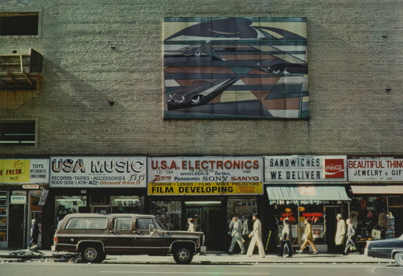 A color photograph of a brick building with colorful signs advertising products for different stores and a large work of art hanging above multiple stores.