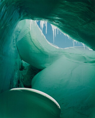 A color photograph from inside of a blue and green ice cave showing a sliver of blue sky.