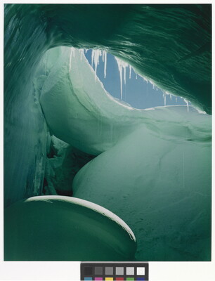 A color photograph from inside of a blue and green ice cave showing a sliver of blue sky.